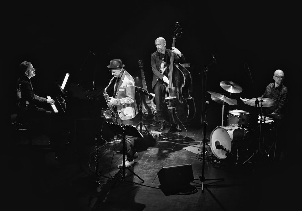 Czarno-białe zdjęcie czteroosobowego zespołu na scenie. Pianista, saksofonista, kontrabasista i perkusista. Tło ciemne.