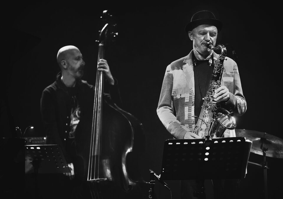 Czarno-białe zdjęcie dwóch muzyków. Po lewej basista z kontrabasem. Po prawej saksofonista. Przed nimi pulpity z nutami. Tło jest ciemne.