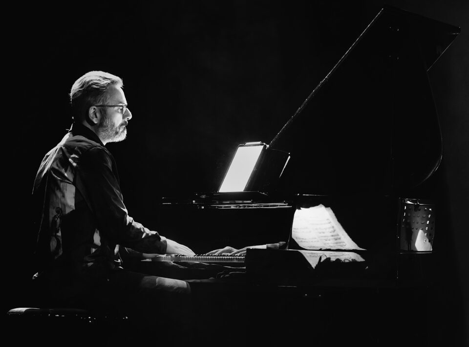 Czarnobiałe zdjęcie mężczyzny grającego na fortepianie w ujęciu z profilu. Mężczyzna ma ciemny strój i okulary. Siedzi przy fortepianie z rękami na klawiaturze. Tło jest czarne.