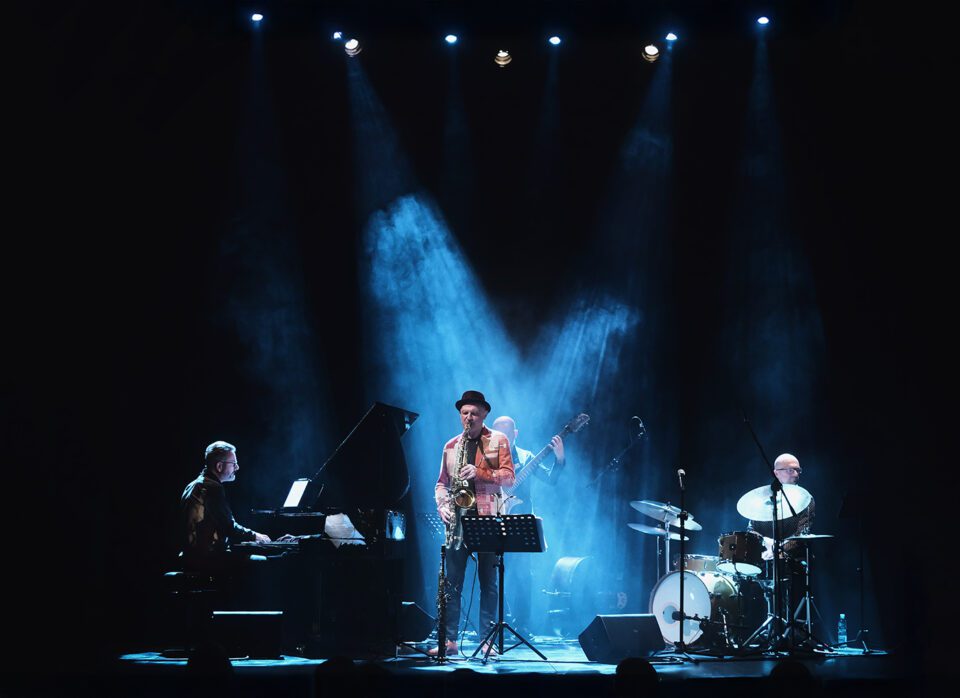 Zdjęcie zespołu muzycznego na scenie. W centrum saksofonista w niebieskawym świetle reflektorów. Za nim basista. Po lewej pianista. Po prawej perkusista. Tło czarne.