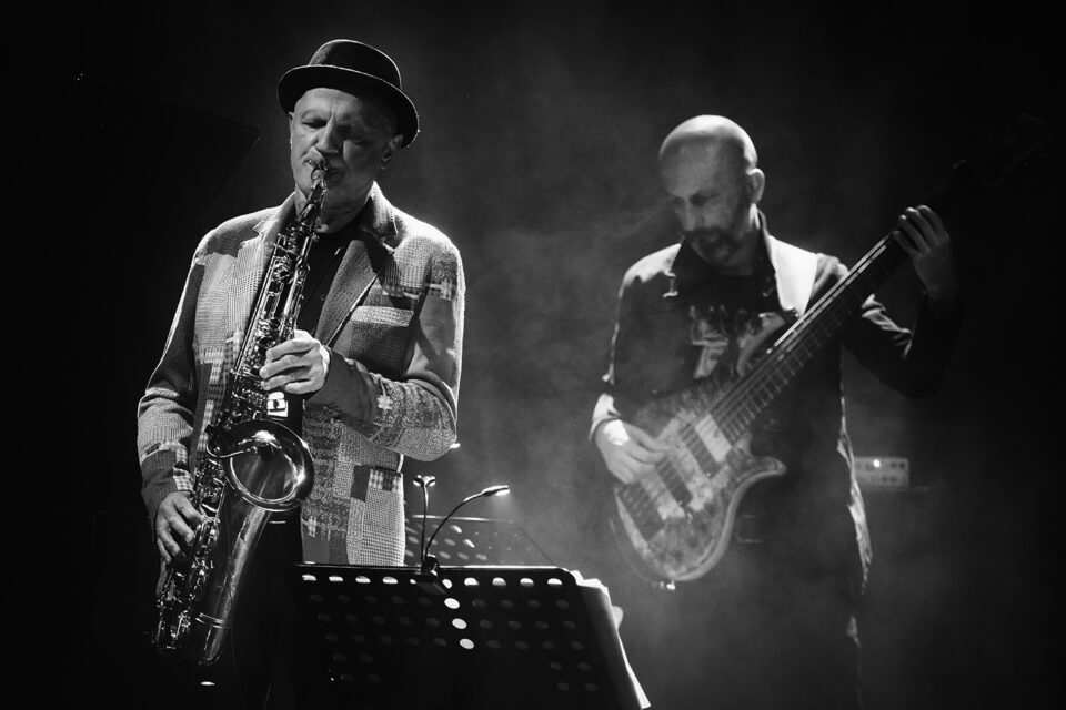 Czarno-białe zdjęcie dwóch grających muzyków. Po lewej saksofonista, po prawej basista. Tło ciemne.