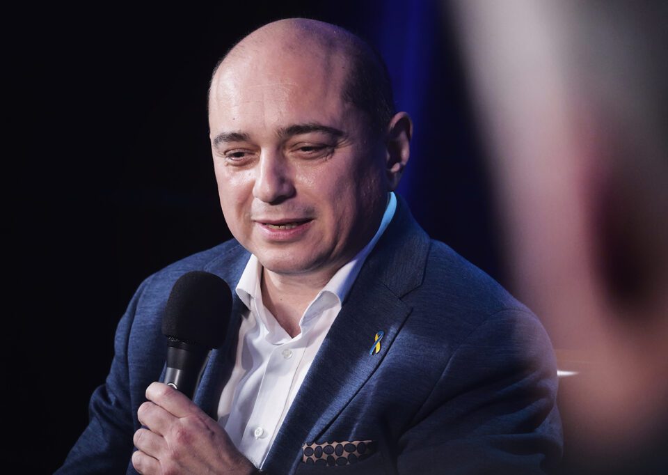 Zdjęcie portretowe uśmiechniętego mężczyzny. Mężczyzna jest łysy, jest ubrany w białą koszulę i granatową marynarkę. Trzyma przed sobą czarny mikrofon. Tło ciemne.