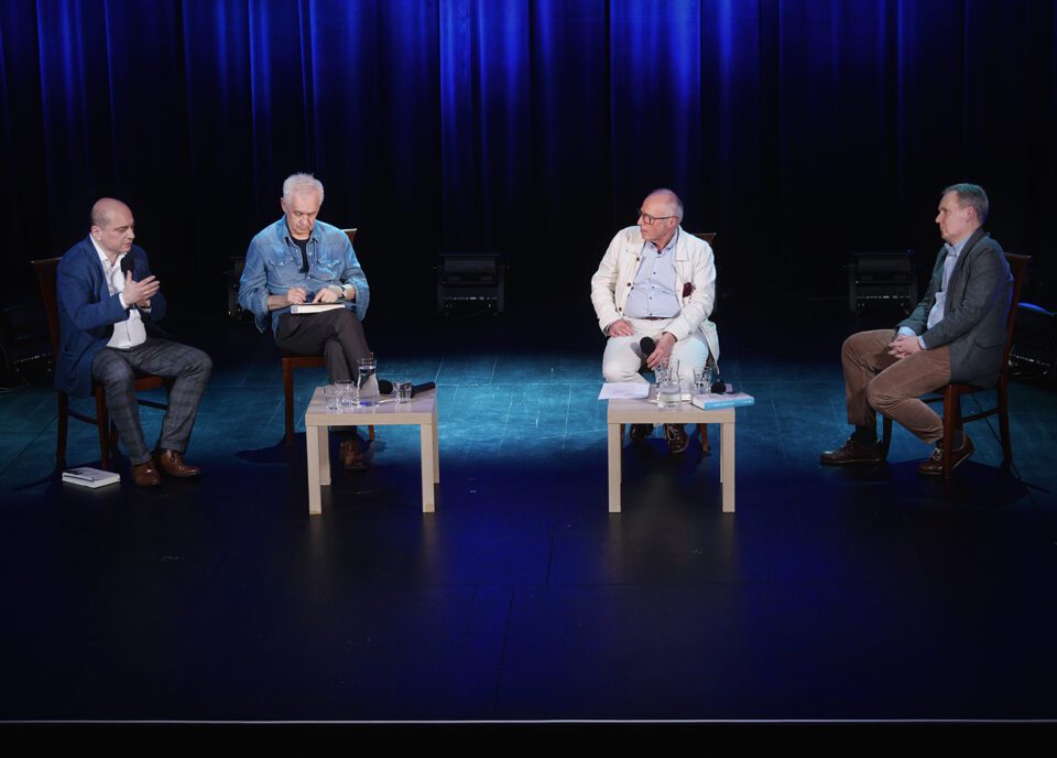 Czterech mężczyzn na scenie. Siedzą na ciemnych krzesłach. Przed nimi dwa, małe, jasne stoliki. Scena ciemna. W głębi sceny kotara oświetlona na niebiesko.