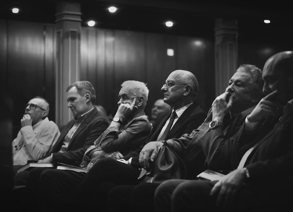 Czarno-białe zdjęcie sześciu mężczyzn siedzących w pierwszym rzędzie w teatrze. Są zamyśleni i elegancko ubrani. W tle sala widowiskowa.