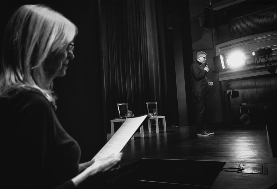 Czarno-białe zdjęcie kobiety i mężczyzny. Ujęcie z boku sceny. Kobieta po lewej w ujęciu z profilu. Trzyma przed sobą kartkę. Ma jasne włosy i ciemny strój. Po prawej w oddali mężczyzna w ciemnym garniturze.