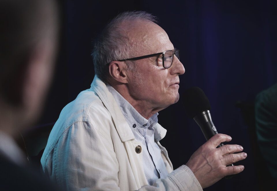 Zdjęcie starszego mężczyzny w ujęciu z profilu. Ma jasną koszulę i jasną kurtkę z kołnierzem. Ma okulary. Trzyma przed sobą mikrofon. Tło jest ciemne.