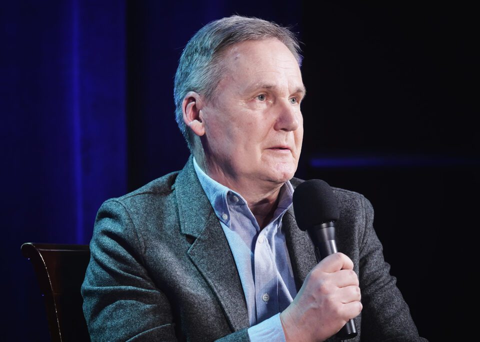 Zdjęcie portretowe siwowłosego mężczyzny. Jest ubrany w błękitna koszulę i szarą marynarkę. Przed sobą trzyma mikrofon. W tle ciemna kotara z refleksami niebieskiego światła.