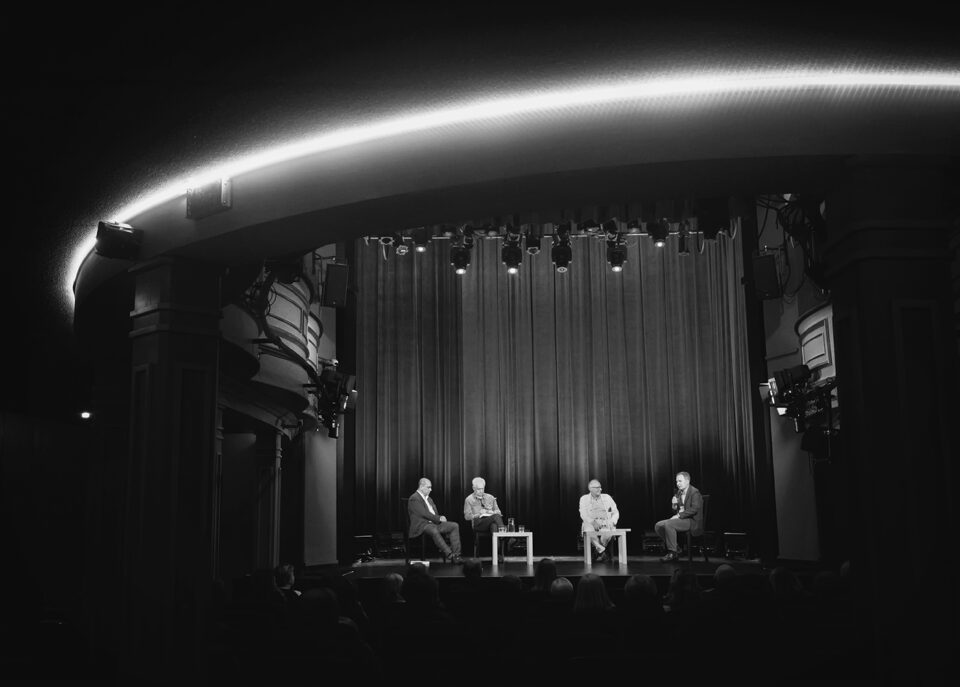 Zdjęcie sali widowiskowej. W głębi ujęcia scena. Na scenie czterech mężczyzn siedzących na krzesłach, za nimi wysoka kotara. U góry świetlisty łuk.