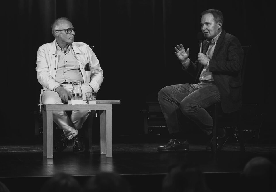 Czarnobiałe zdjęcie dwóch mężczyzn siedzących na krzesłach. Mężczyzna po lewej ma jasny strój. Przed nim stoi mały stolik. Mężczyzna po prawej ma ciemny strój. W jednej ręce trzyma mikrofon, drugą gestykuluje. Tło ciemne.