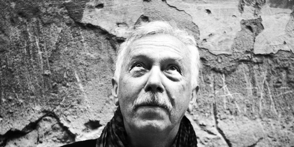 Slajd: Czarno-białe zdjęcie portretowe uśmiechniętego starszego mężczyzny. Ma niemal białe krótkie włosy i wąsy. Pod szyją ma zawinięty ciemny szalik. W tle odrapana ściana z odpadającym tynkiem.