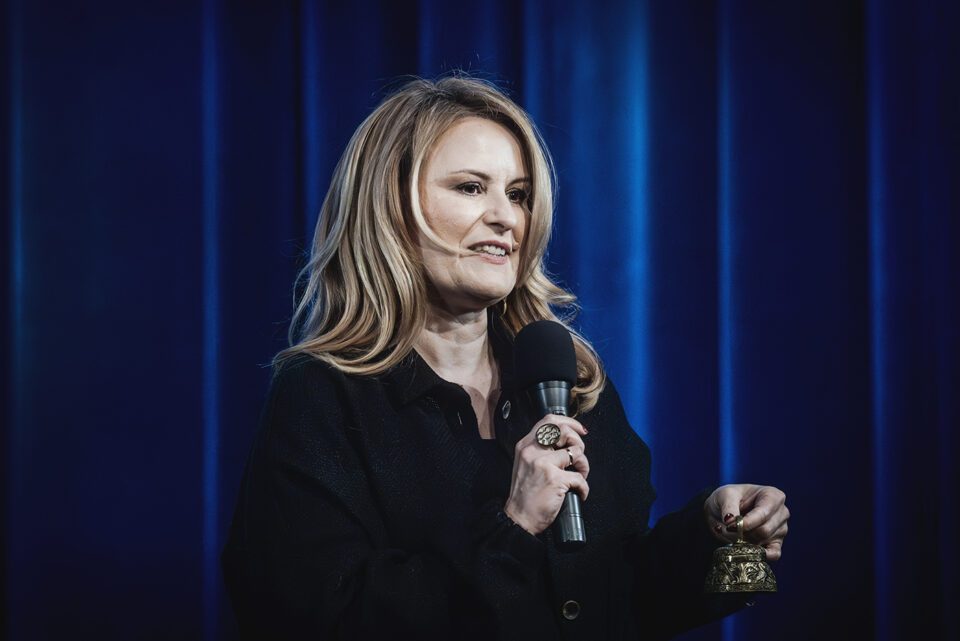 Zdjęcie kobiety w ujęciu od pasa w górę. Kobieta ma jasne, rozpuszczone włosy. Jest ubrana na czarno. W jednej ręce trzyma mikrofon, w drugiej ozdobny dzwoneczek. W tle kotara oświetlona na niebiesko.