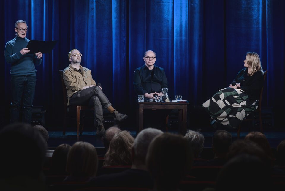 Ujęcie sceny zza pleców widzów. Na scenie są cztery osoby. Prawą i środkową część zajmują trzy osoby siedzące na krzesłach. Przed nimi stoi stolik. Po lewej stoi lektor, trzyma przed sobą tekst na czarnej podkładce. W tle kotara oświetlona na niebiesko.