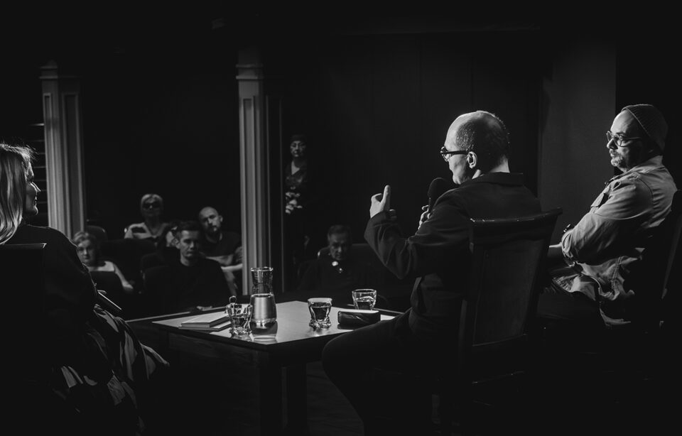 Ujęcie zza pleców gości siedzących na krzesłach na scenie. Po lewej stronie kobieta, po prawej dwóch mężczyzn. Po środku stolik, a na nim książki, karafka i szklanki z wodą oraz mikrofony. W głębi ujęcia widownia teatru. Jasne kolumny i rzędy foteli zajęte przez widzów. Tło ciemne.