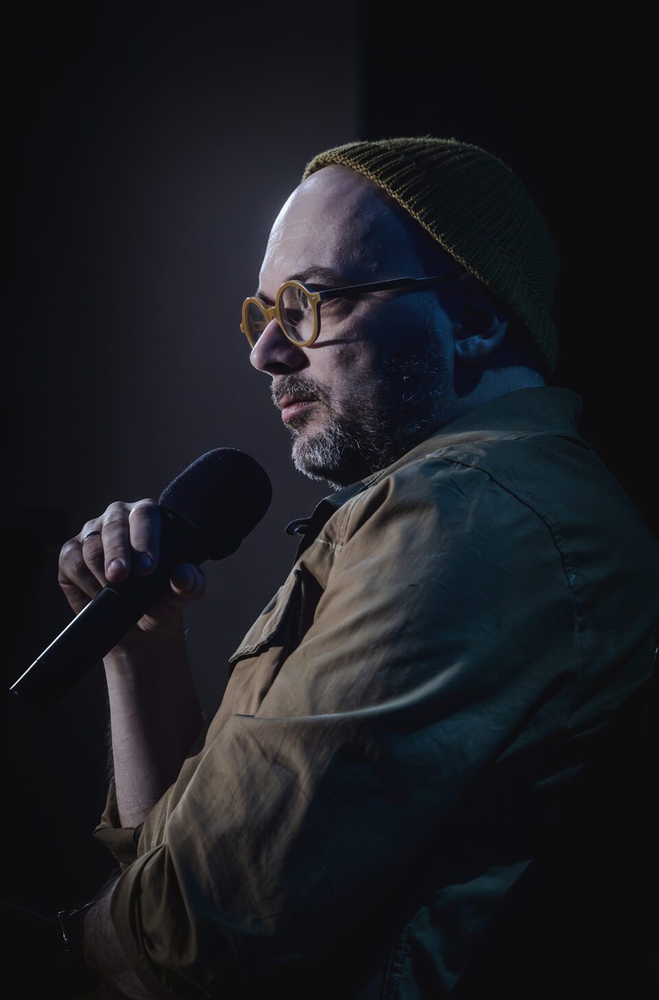 Zdjęcie portretowe mężczyzny w ujęciu z profilu. Mężczyzna trzyma mikrofon. Ma dopasowane kolorystycznie elementy garderoby. Koszulę, czapkę i oprawki okularów w kolorze jasnobrązowym. Tło ciemne.
