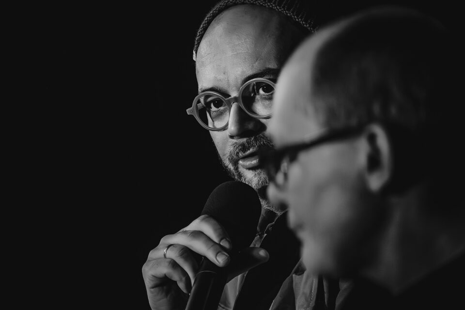 Twarze dwóch mężczyzn w okularach. Mężczyzna na pierwszym planie w ujęciu z profilu jest nieco rozmazany. Ostrość zdjęcia skupia się na drugim mężczyźnie, który trzyma mikrofon przy ustach. Tło czarne.