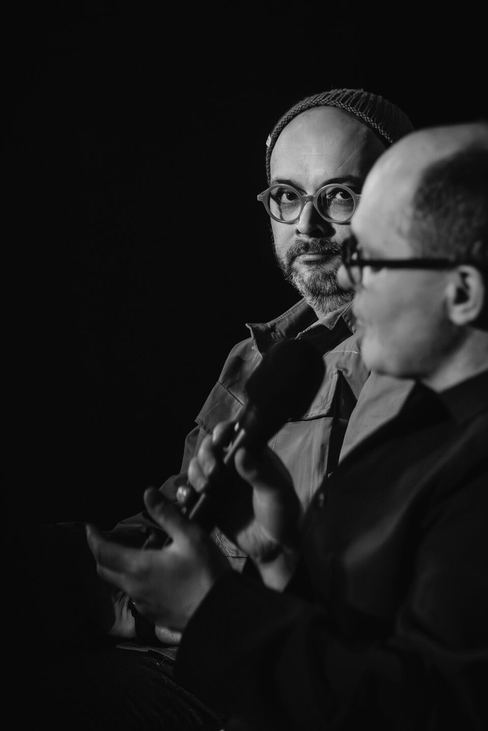 Czarno-białe zdjęcie dwóch mężczyzn. Jeden z nich po prawej w ujęciu z profilu. Dugi jest przez niego częściowo przysłonięty. Obaj są w okularach. Lewą cześć zdjęcia wypełnia czarne tło.