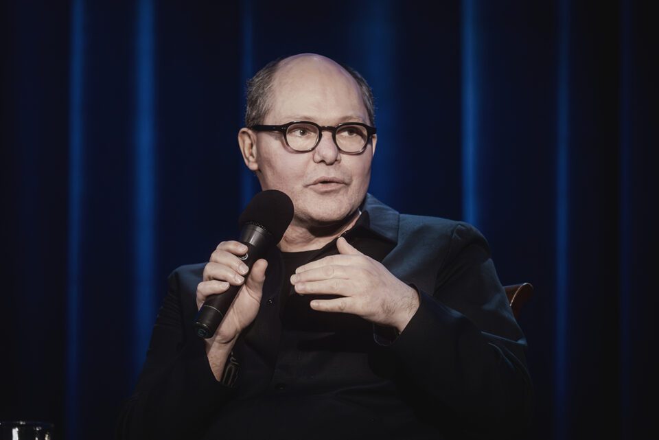 Zdjęcie portretowe mężczyzny w okularach i ciemnym stroju. Trzyma prze sobą mikrofon. W tle ciemna kotara w niebieskimi refleksami.