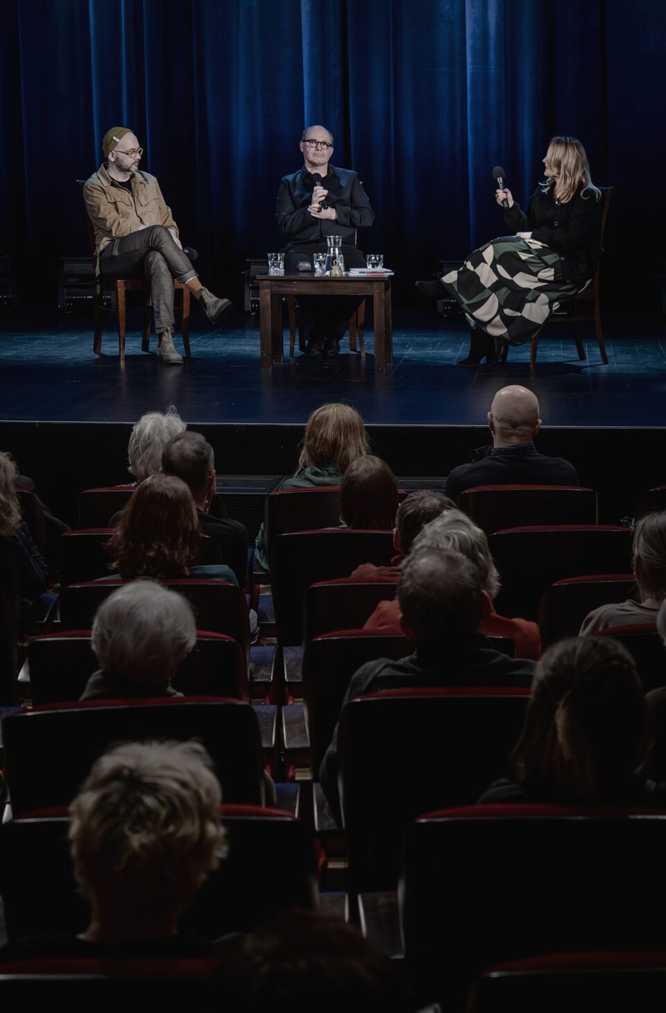 Zdjęcie sceny w ujęciu zza pleców widzów z kilku pierwszych rzędów. Na scenie są trzy osoby siedzące na krzesłach, przed nimi po środku stoi stolik. W głębi sceny ciemna kotara z niebieskimi refleksami.