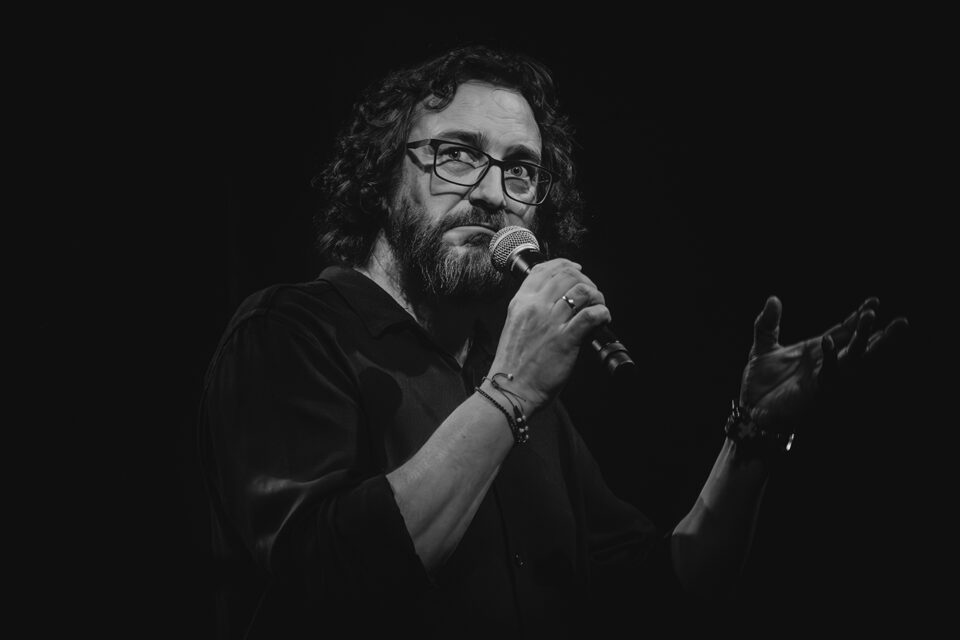 Zdjęcie mężczyzny w ujęciu od pasa w górę. Ma kręcone włosy, brodę i wąsy. Jest w okularach i ciemnym stroju. Tło czarne.