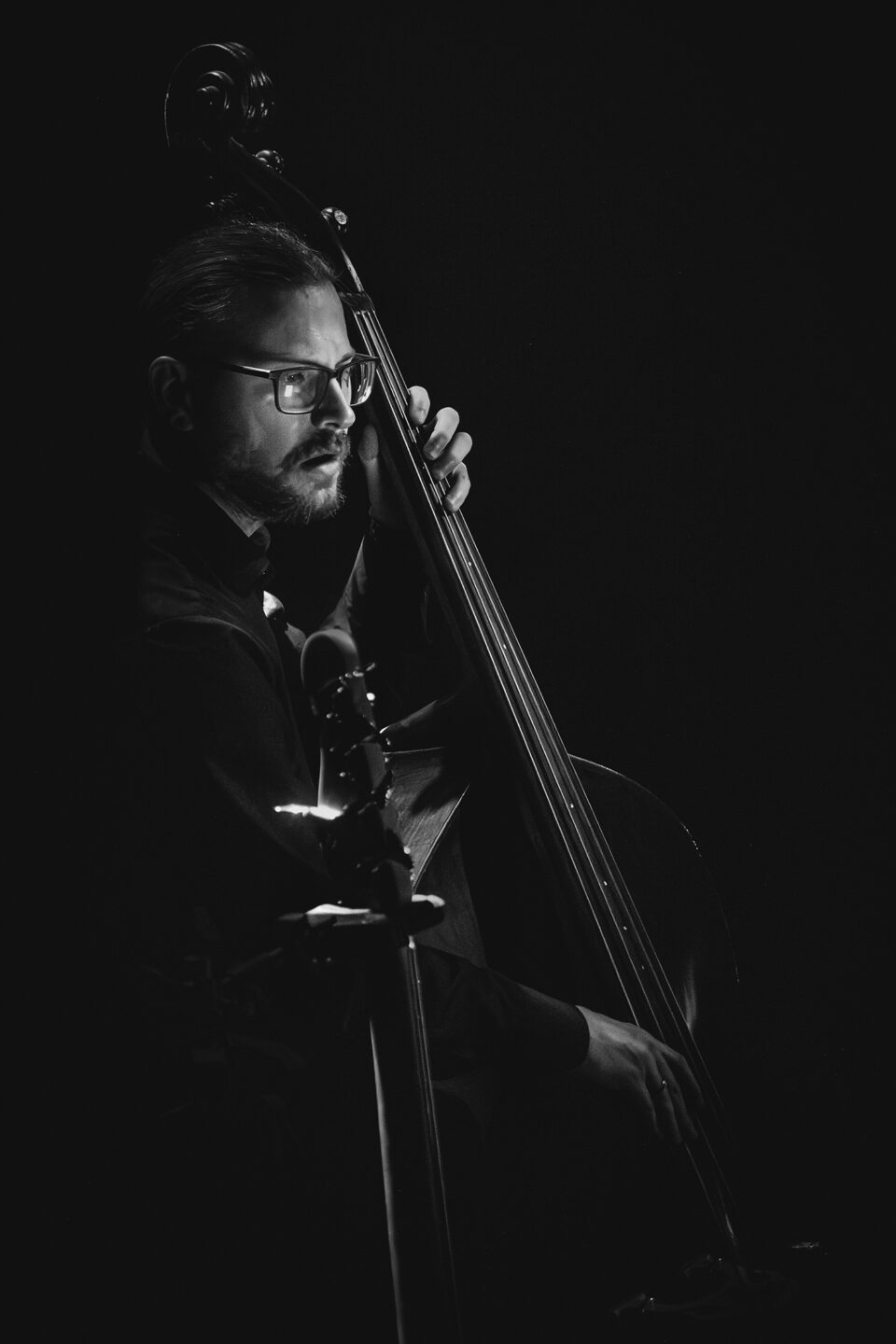Czarno-białe zdjęcie mężczyzny grającego na kontrabasie. Mężczyzna ma ciemne włosy, brodę i wąsy. Jest w ciemnym stroju i okularach. Tło ciemne.
