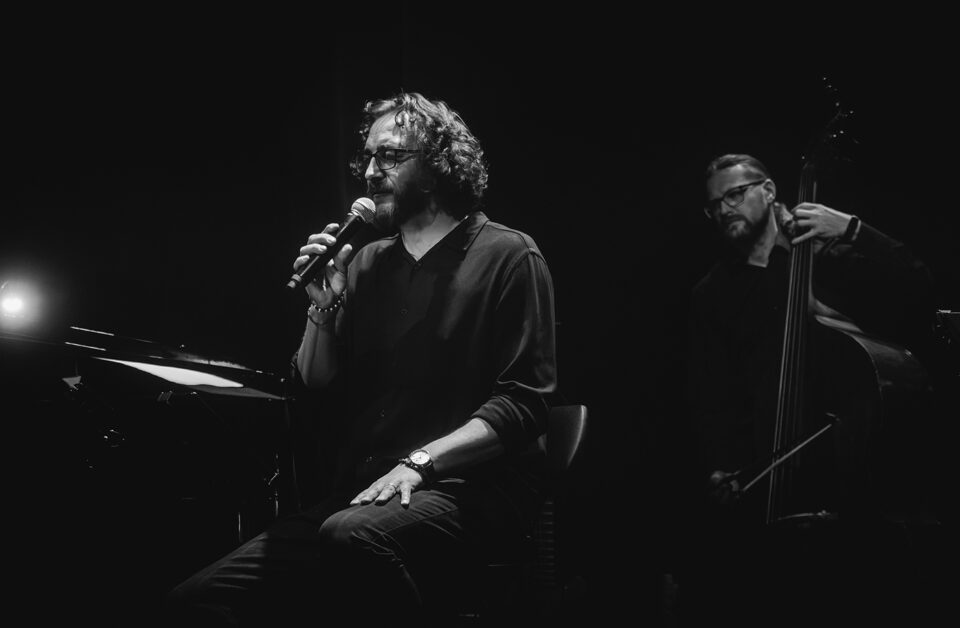 Czarno-białe zdjęcie dwóch muzyków. Na pierwszym planie po lewej wokalista w ujęciu z pół profilu. Siedzi na wysokim stołku, trzyma mikrofon. Na drugim planie po prawej Kontrabasista. Tło czarne.