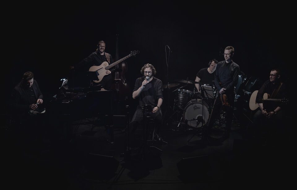Sześcioosobowy zespół muzyczny na scenie. Mężczyźni ubrani w ciemne stroje. Pianista, basista, wokalista, perkusista, skrzypek i gitarzysta. Tło ciemne.