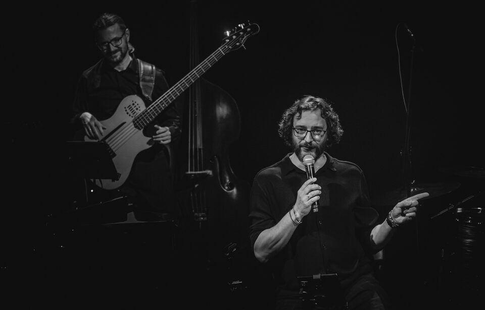 Czarno-białe zdjęcie dwóch muzyków w ciemnych strojach. Na pierwszym planie po prawej wokalista z mikrofonem. Ma okulary, kręcone włosy sięgające za ucho oraz brodę i wąsy. Na drugim planie po lewej basista z gitarą basową. Ma okulary, brodę i wąsy. Tło czarne.