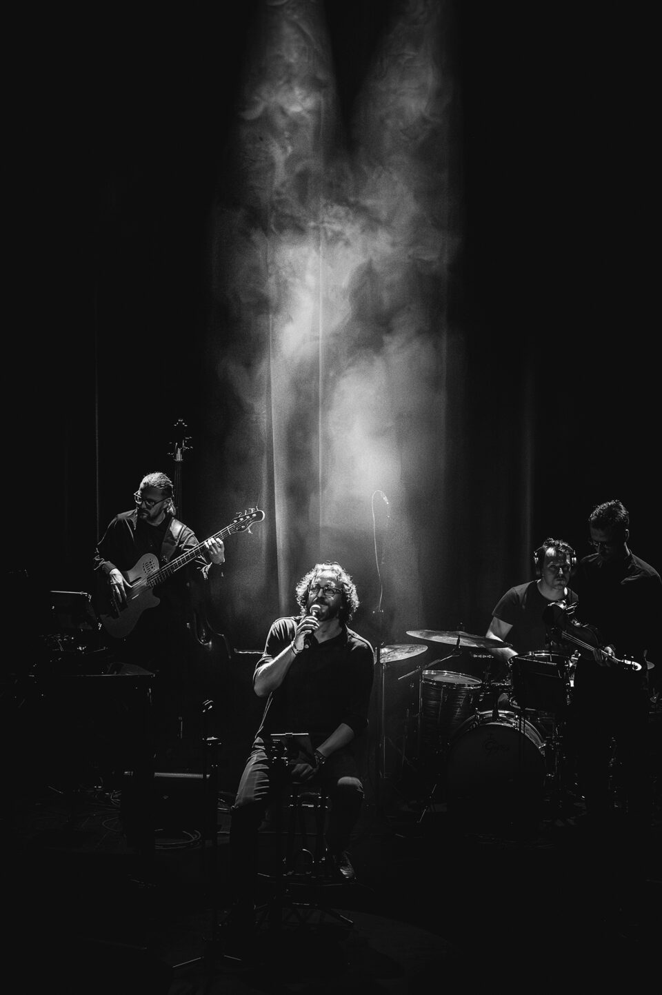 Czarno-białe zdjęcie czterech muzyków na scenie. Na pierwszym planie wokalista siedzący na scenie, tuż za nim snop jasnego światła rozproszonego w sztucznym dymie. Na drugim planie basista, perkusista i skrzypek. Tło ciemne.