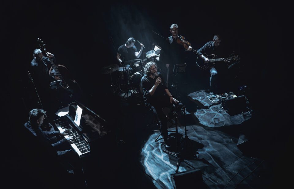 ujęcie sceny z góry. na scenie sześciu muzyków. W pierwszym rzędzie pianista, wokalista, i gitarzysta. W drugim rzędzie kontrabasista, perkusista i skrzypek. Tło ciemne z niebieskawym refleksami światła.