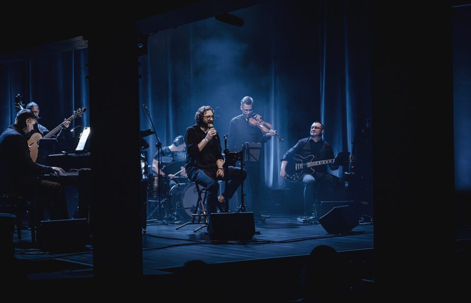 Zdjęcie sześcioosobowego zespołu muzycznego na scenie. Światło niebieskawe rozproszone w delikatnym sztucznym dymie. W tle ciemna kotara z niebieskimi refleksami.