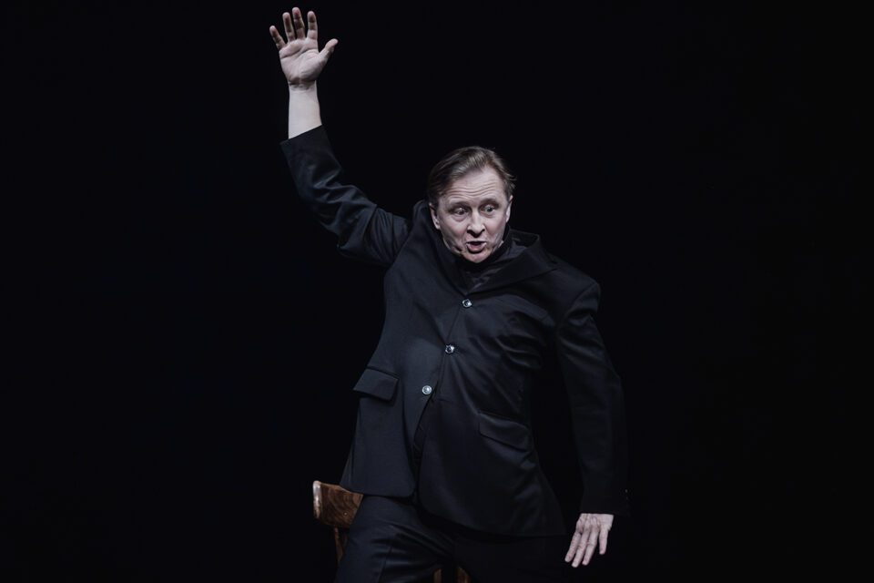 Zdjęcie mężczyzny w czarnym stroju. W ekspresyjnej pozie. Jedną rękę ma podniesioną wysoko z rozłożonymi palcami, drugą trzyma bardzo nisko, jakby chciał pokazać nią coś małego. Tło ciemne.