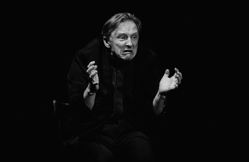 Mężczyzna w czarnym stroju. Ma minę wyrażającą rozpacz i złość. Trzyma przed sobą ręce z lekko ugiętymi palcami, uniesione na wysokości klatki piersiowej. Tło czarne.