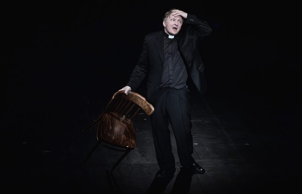 Mężczyzna stojący na scenie. Jedną ręką trzyma oparcie przechylonego krzesła. Drugą ręką ociera czoło. Jest ubrany w czarną koszulę z koloratką, czarne spodnie i marynarkę. Tło ciemne.