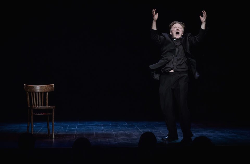 Zdjęcie mężczyzny w czarnym stroju na scenie. Po lewej stoi drewniane krzesło. Po prawej mężczyzna z rękami uniesionymi do góry. Ma ekspresyjną pozę i minę z szeroko otwartymi ustami. Na deski sceny pada delikatne światło. W tle ciemność.