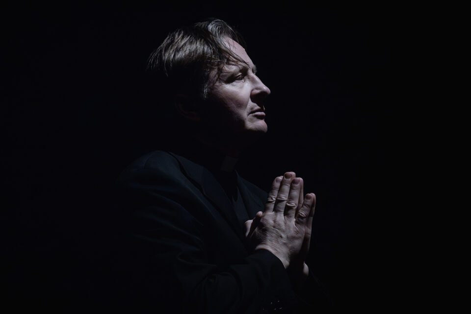 Zdjęcie mężczyzny w ujęciu z profilu. Jest ubrany na czarno. Na wysokości klatki piersiowej ma dłonie złożone do modlitwy. Ma poważną minę. Tło jest czarne.