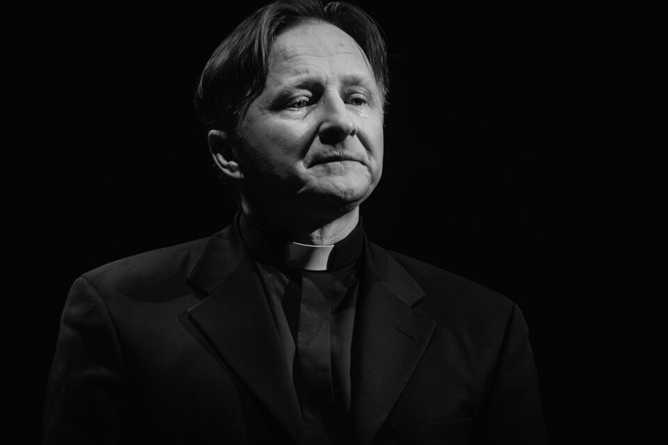 Zdjęcie portretowe mężczyzny w czarnym stroju z koloratką. Mężczyzna ma poważną, spokojną minę. Tło czarne.