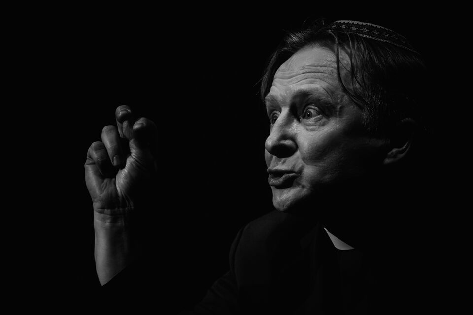 Czarno-białe zdjęcie twarzy mężczyzny w ujęciu z półprofilu. Mężczyzna ma szeroko otwarte oczy i układa usta w dziubek. Prawą dłoń trzyma z zaciśniętymi palcami na wysokości twarzy. Tło czarne.