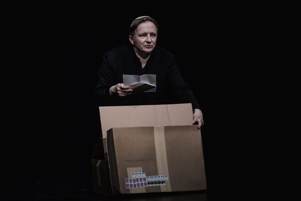 Mężczyzna w czarnym stroju. Przed nim duże tekturowe pudełko z naklejonymi znaczkami pocztowymi. Mężczyzna jedną ręką dotyka pudełka, w drugiej, ponad pudełkiem, trzyma list. Tło czarne.