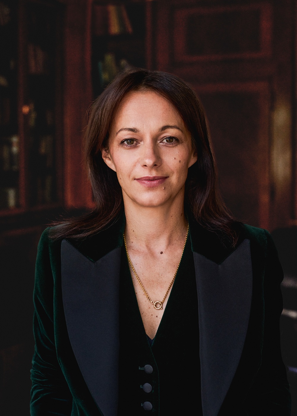 Zdjęcie portretowe uśmiechniętej kobiety. Jest ubrana w czarną bluzkę z trójkątnym dekoltem i ciemny żakiet. W rozmytym tle wnętrze ciemnego pomieszczenia.