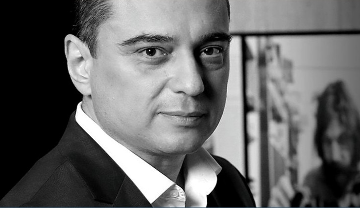 Czarno-białe zdjęcie portretowe mężczyzny. Ma ciemne włosy, jest ubrany w białą koszulę i czarną marynarkę. Tło po lewej czarne, po prawej w rozmytym tle jest wnętrze pomieszczenia z obrazem mężczyzny z brodą.