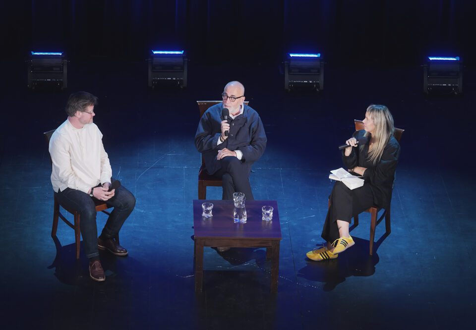 Ujęcie na troje rozmówców, zdjęcie wykonano znad sceny. Niebieskawe światło.