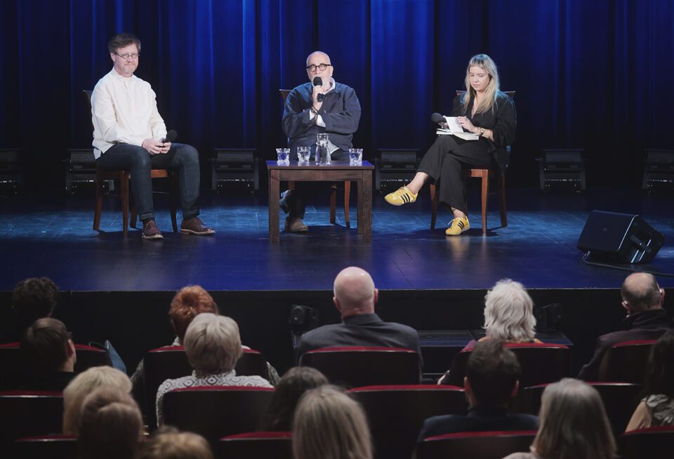 Zdjęcie zrobione z perspektywy widowni; na scenie wszyscy rozmówcy: Laskowski, Kwieciński i prowadząca spotkanie Maja Chitro; wszyscy siedzą na krzesłach przy stoliku z postawioną wodą. W tle kotara oświetlona na niebiesko.