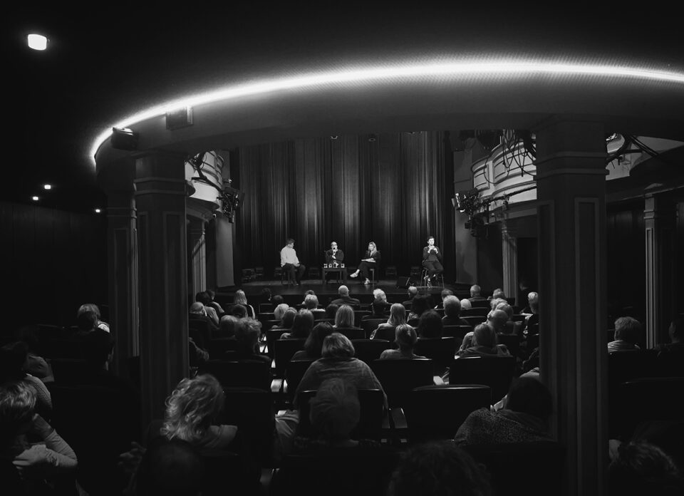Zdjęcie zrobione z tylnych rzędów widowni, czarno-białe; na pierwszym planie gęsto obsadzone fotele; w tle rozmówcy i obok nich - tłumaczka PJM.