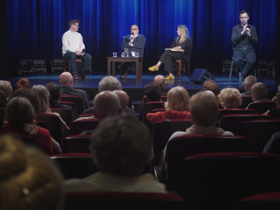 Ujęcie sceny zza pleców widzów. Na scenie rozmówcy i tłumacz PJM. W tle niebieska kotara.