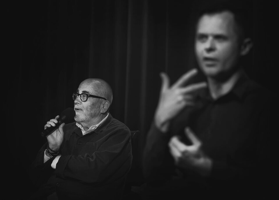 Czarno-biały portret Michała Kwiecińskiego; na pierwszym planie rozmyta sylwetka tłumacza PJM.