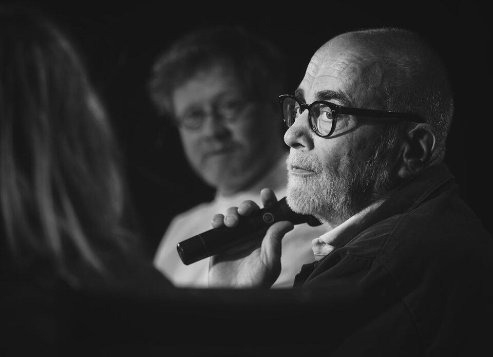 Czarno-białe zdjęcie, wąski kadr, w centrum twarz Michała Kwiecińskiego, ten spogląda na Maję Chitro, której głowa, uchwycona od tyłu, jest na pierwszym planie, rozmyta; w tyle rozmyta twarz Aleksandra Laskowskiego.