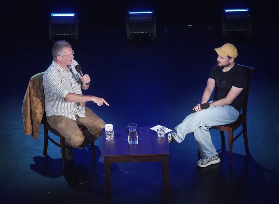 Kadr z góry, Meller i Szot rozmawiają, Meller stanowczym gestem z wyprostowanym palcem wskazującym nakierowanym na podłogę podkreśla wagę swoich słów.