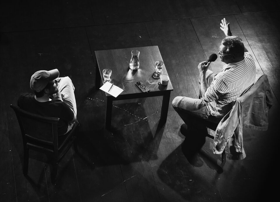 Zdjęcie wykonane nad głowami rozmówców; mężczyźni rozmawiają, Meller gestykuluje, pomiędzy nimi stolik - na nim karafki z wodą, telefon jednego z nich i notatki Szota.