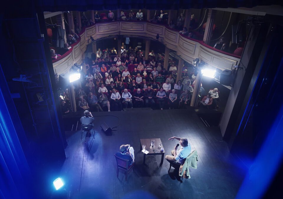 Widok na scenę i widownię; niemal wszystkie miejsca są zajęte; dolna część kadru jest oświetlona na niebiesko - to dekoracje kotary.