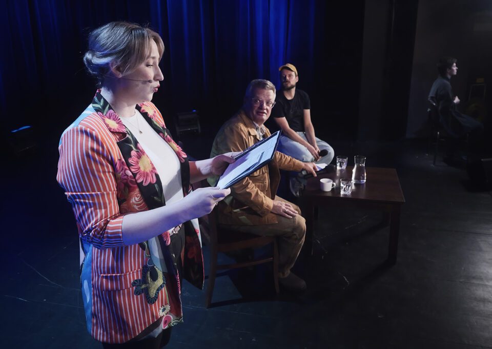 Pierwsze z cyklu 12 zdjęć; zrobiono je na scenie teatru; na pierwszym planie aktorka czytająca fragment książki Marcina Mellera, za nią autor i prowadzący spotkanie - Wojtek Szot, w tle tłumaczka języka migowego.